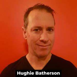 A white man with sshort brown hair and a black shirt in front of an orange wall. The man smiles at the camera. At the bottom of the photo, in white text on a black rectangle, is the words 'Hughie Batherson'.