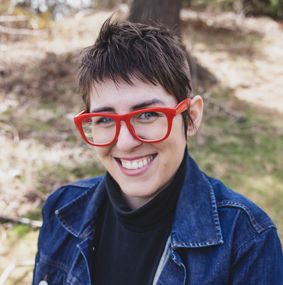 A white nonbinary person with short brown choppy straight hair, bright red large framed glasses, wearing a black turtleneck and blue denim jean jacket, sitting outside smiling at the camera.